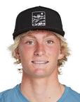 Headshot of a young man wearing a black hat with a white, rectangular framed print of choppy water with the letters I P D above the water and a circular sun in the top right corner of the frame.