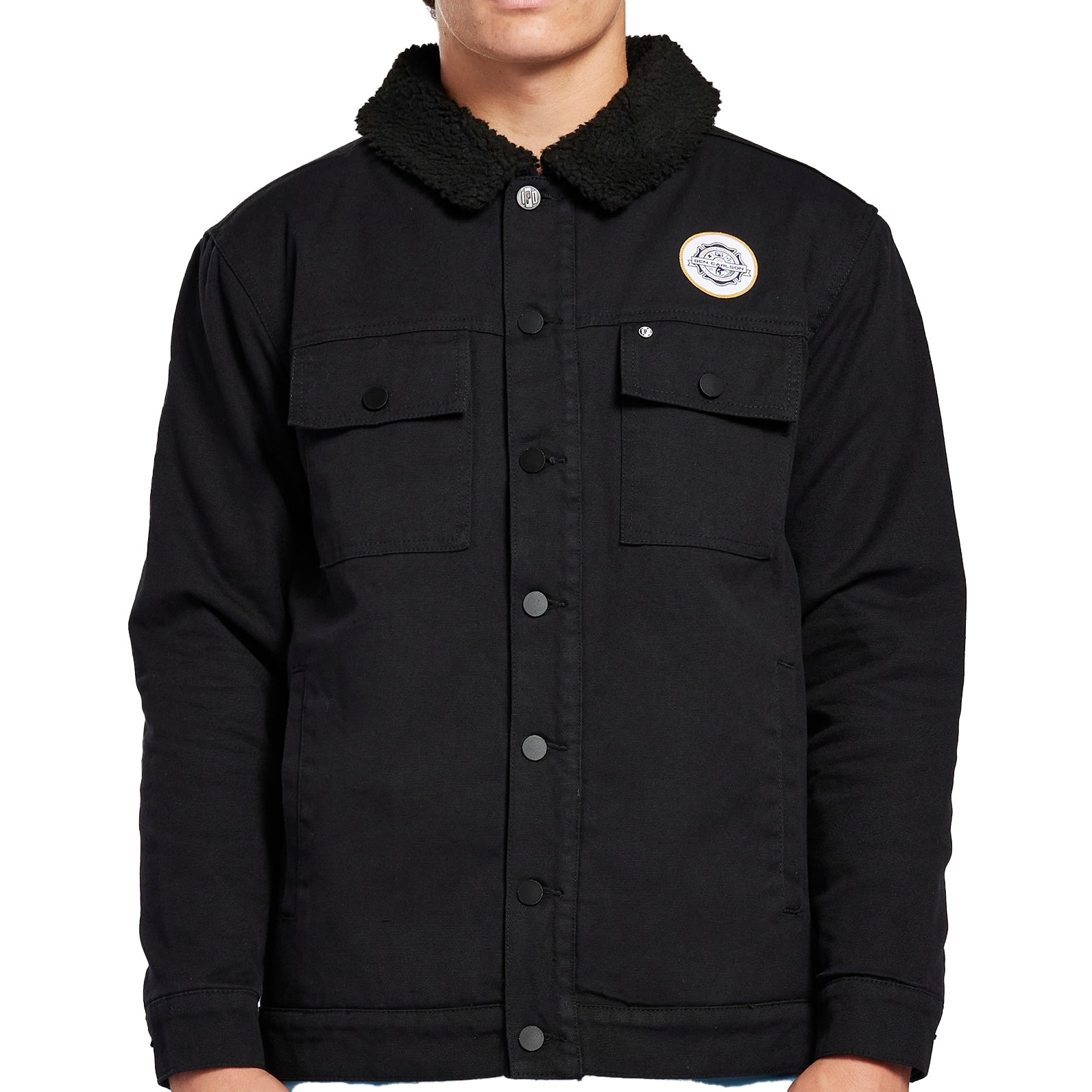 Front view of a black long sleeve heavyweight cotton button up jacket. It has a thick, cotton-lined collar with two patch flap pockets on each side of the chest. Above the left pocket is a white circular patch.
