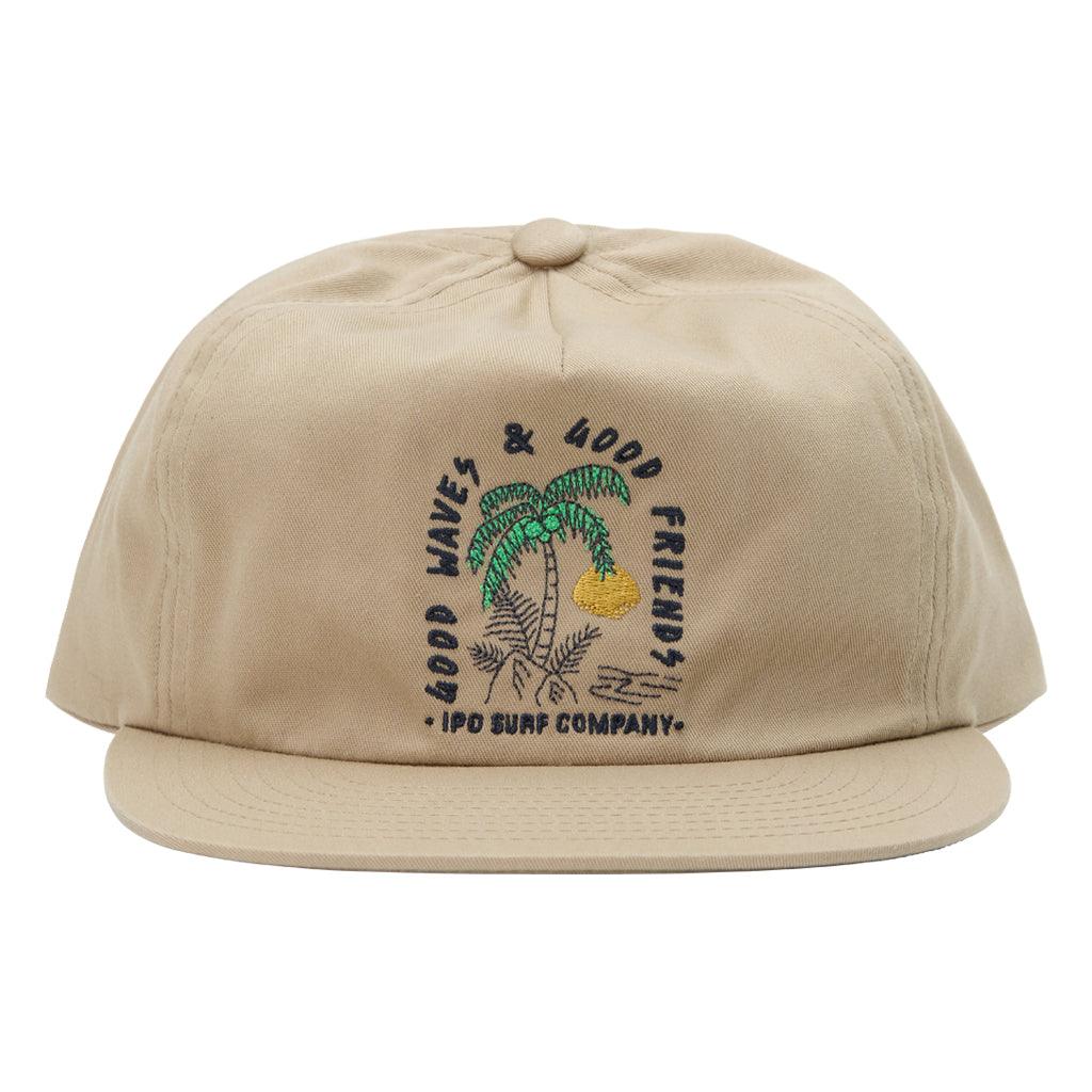 Front view of a tan hat with a print of a palm tree and fern growing behind a rock on a beach with the setting sun in the background. Arching around the image are the words Good Waves and Good Friends. Below the image are the words I P D Surf Company.
