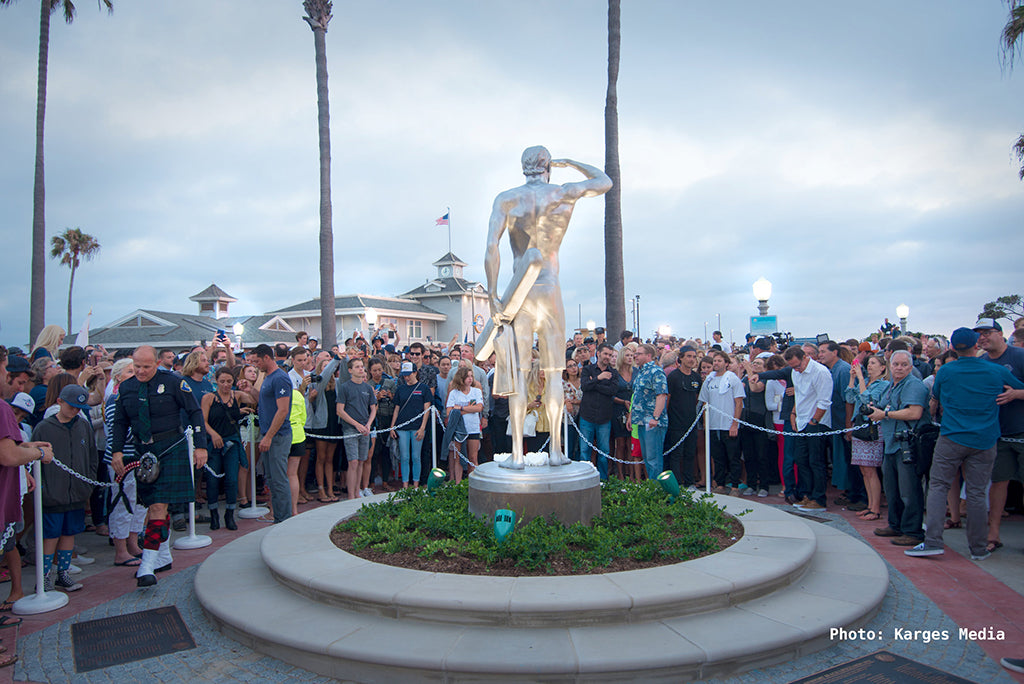 Rear view of a crowd of people outside admiring a large silver statue of a lifeguard. The lifeguard is holding a rescue tube and fins in his left hand and holding his right hand over his eyes while gazing out into the horizon.
