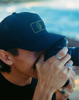 black 1697 trucker hat worn by a person taking photo with camera held to his face. 
