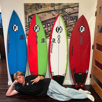 asher posing lying down on one side in front of a colorful collection of 4 surfboards