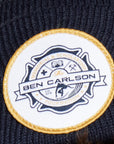 White circular patch with gold trim and a fire department-style badge in the center. Across the badge on a banner are the words The Ben Carlson. Arching above the banner are the words Memorial and Scholarship. Arching below the banner is the word Foundation with two stars on each side. There's a small image of a surfer crouching and riding a wave on his surfboard below the banner.