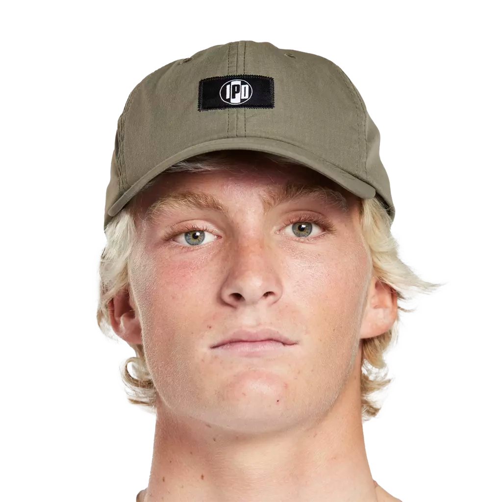 Headshot of a young man wearing an olive green hat with a small black rectangular patch on the front. In the middle of the patch is the I P D logo.