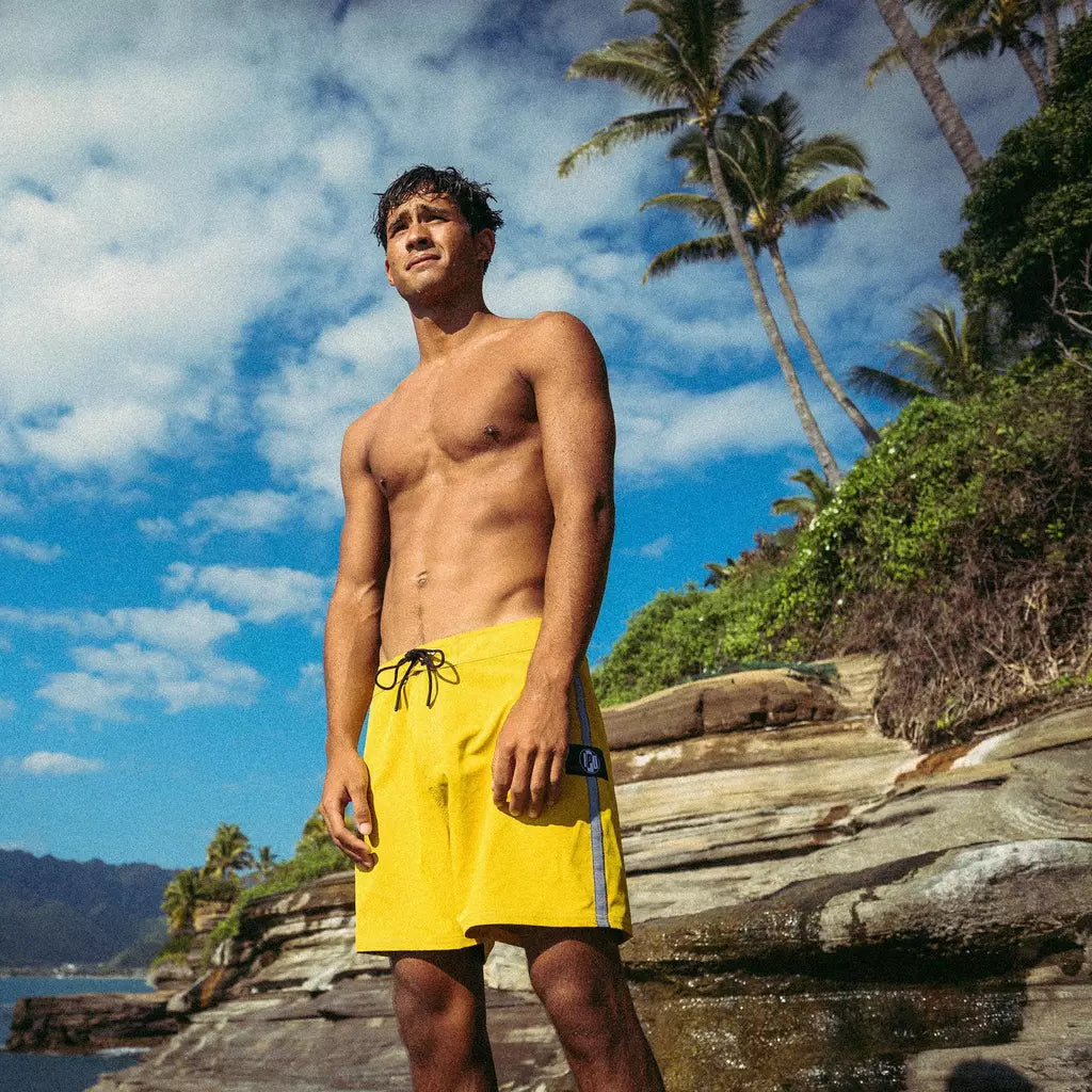 A shirtless man wearing yellow boardshorts standing on the edge of a rocky tropical coastline.