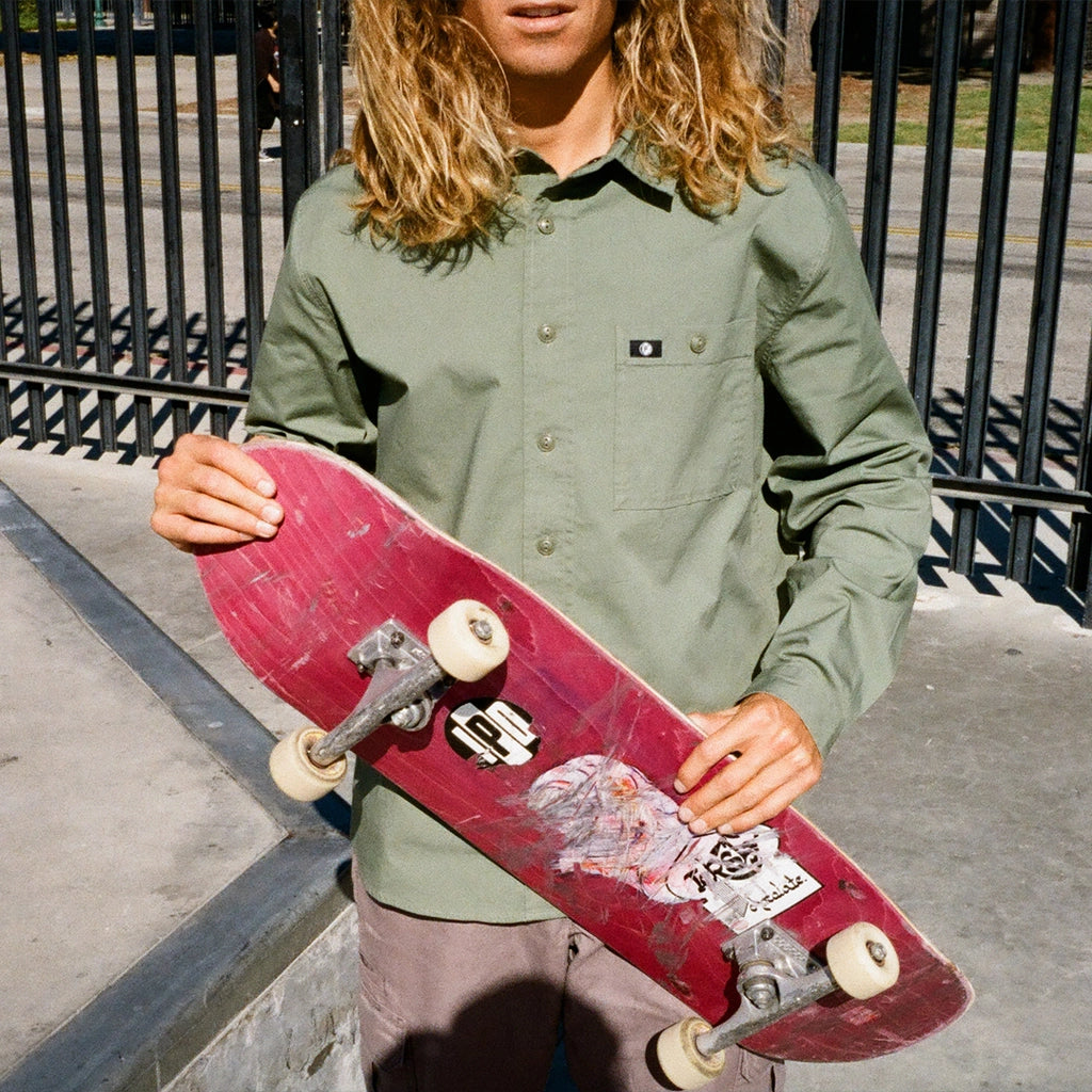 A skater wearing a moss green canvas button down shirt holds up his skateboard.