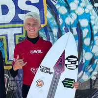 A young surfer posing for the camera while holding his surfboard in his right hand and throwing up a shaka sign with his left hand.