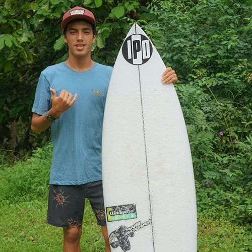 donovan posing next to his surfboard outside, looking stoked, and throwing up a shaka sign