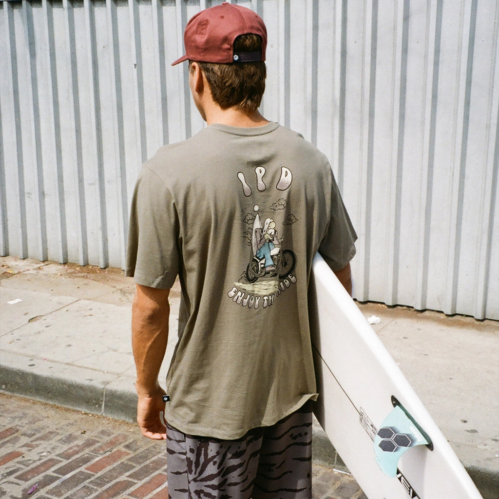 A surfer carries his surfboard while wearing an olive tee with I P D letters and image on the back