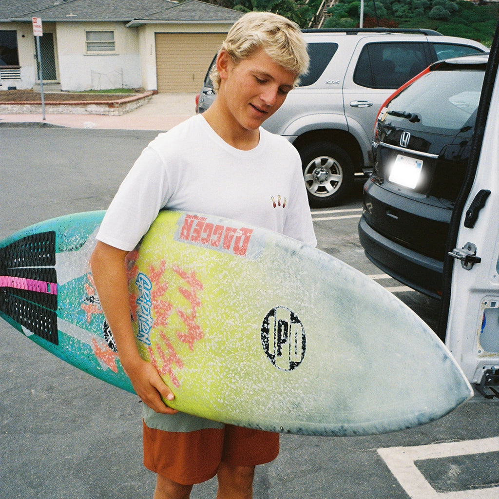 A surfer holding his surfboard while wearing a white tee with I P D letters on the front.