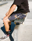 A skater sits on his skateboard while wearing black boardshorts with a floral pattern.