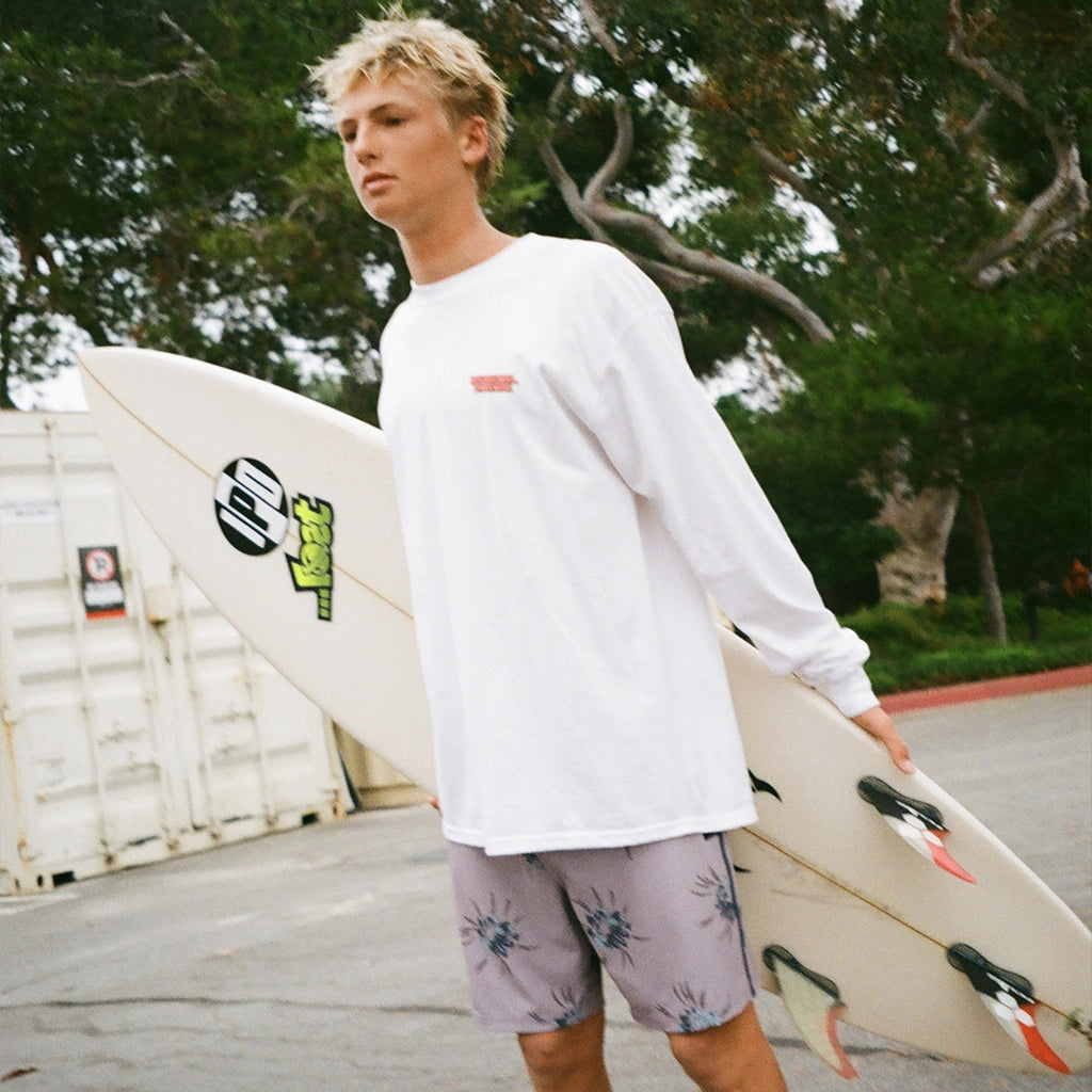 A surfer holds his surfboard while wearing gray moonstone boardshorts with a floral pattern.