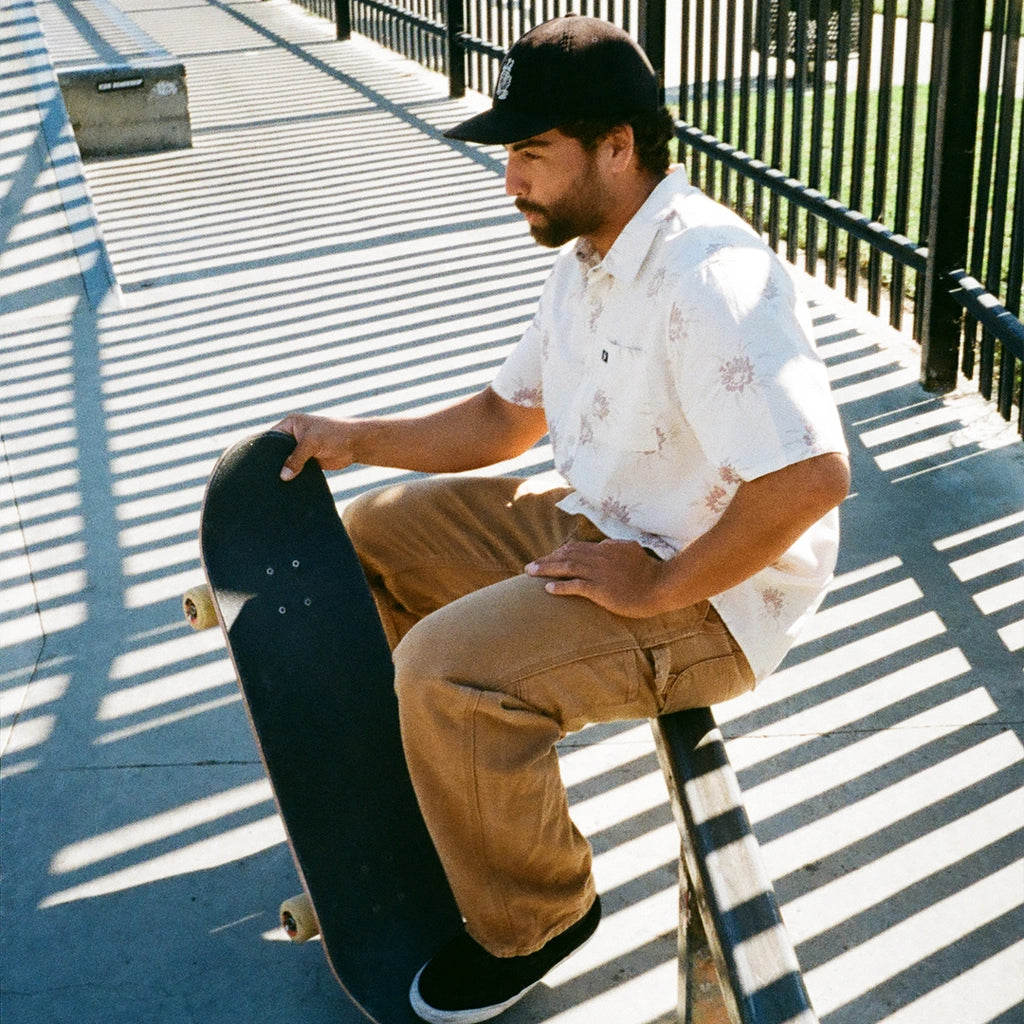 A skater sits on a rail while wearing a white haze short sleeve button up shirt with a floral pattern.