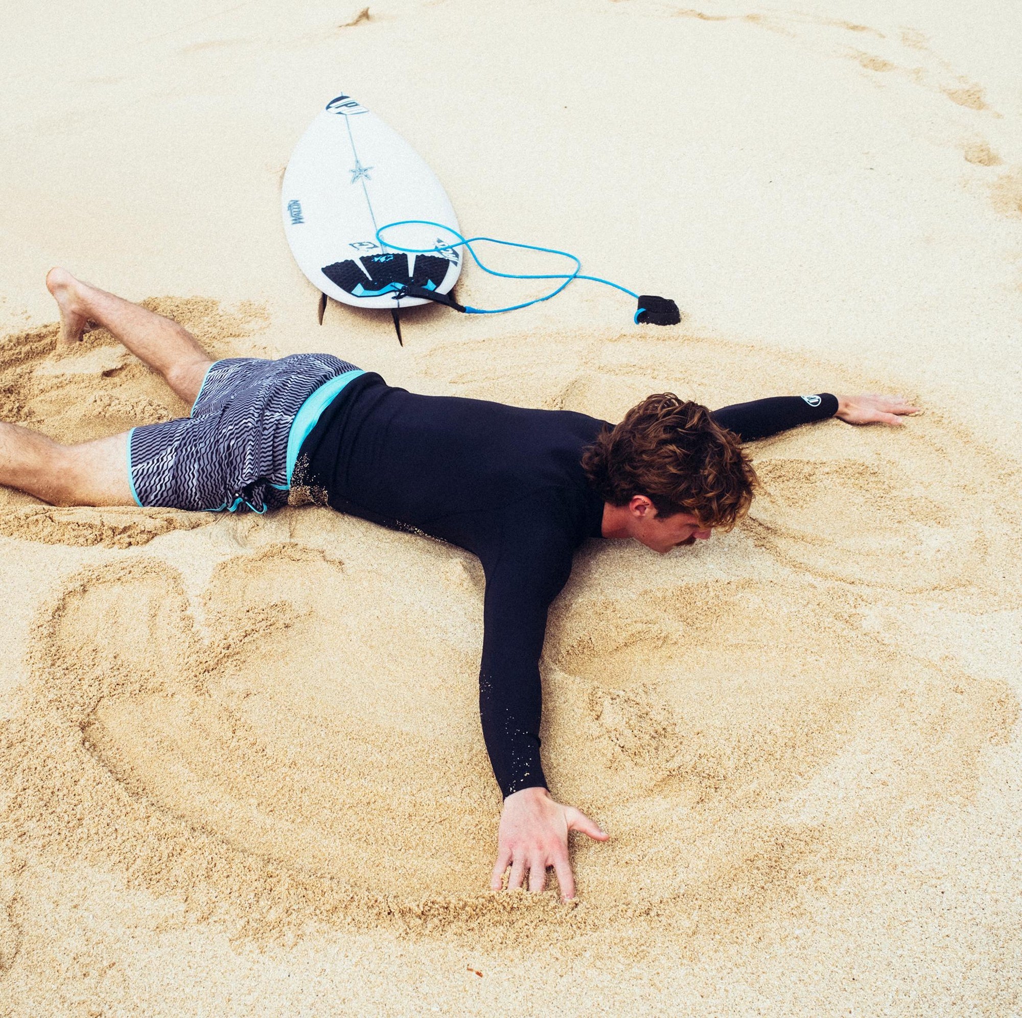 Young man laying face down in the sand making a sand angel while wearing black and silver abstract striped boardshorts mimicking sound frequency with a turquoise trim.