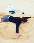 Young man laying face down in the sand making a sand angel while wearing black and silver abstract striped boardshorts mimicking sound frequency with a turquoise trim.