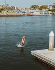 Young man diving into the water mid-dive with his torso in the water wearing dark blue, olive green, and red abstract striped boardshorts mimicking sound frequency.