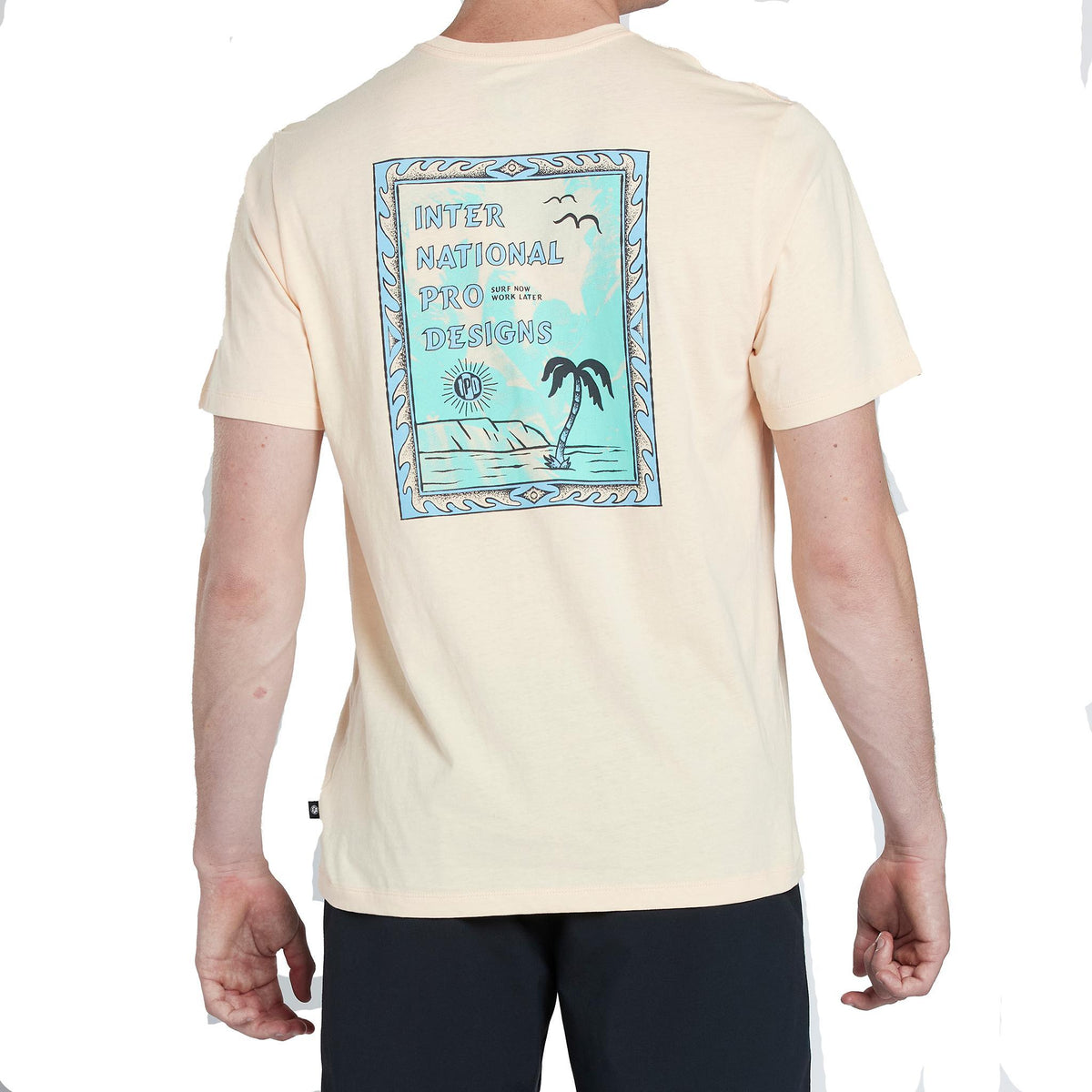 modeling showing the rear view of the mens framed super soft s/s tee in sherbert. showing the large rear graphic of stamp with international pro designs by a palm tree