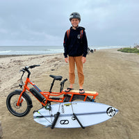 grey smiling and standing on the back of his e-bike on the beach with surfboard attached