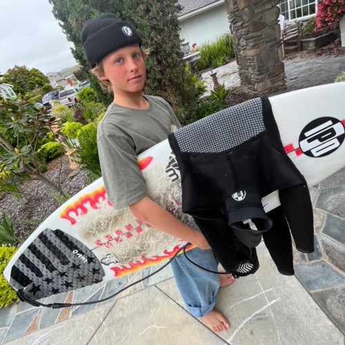 hudson looking content, holding his surfboard in the front yard, wetsuit hanging off the side