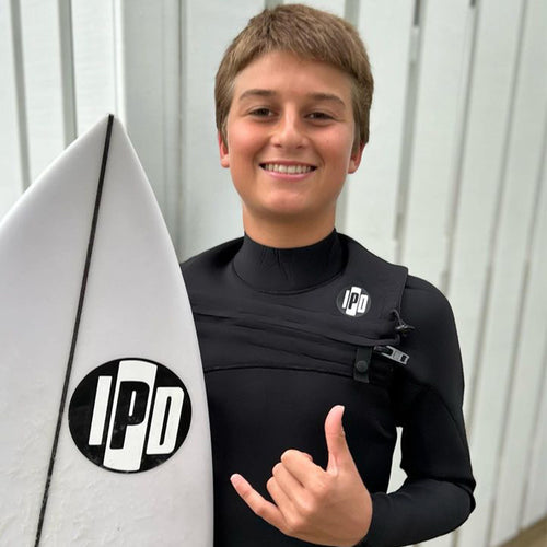 jack standing with his surfboard with an ipd wetsuit on throwing up a shaka sign with a big smile on his face