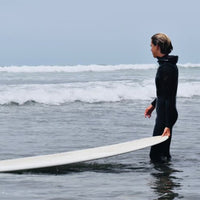 jude grabbing his board in the water with a wetsuit on, looking out at the waves and assessing the conditions