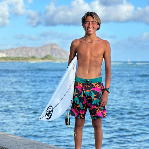 kielan standing in front of a beautiful hawaiian ocean view, holding his surfboard and wearing some colorful board shorts