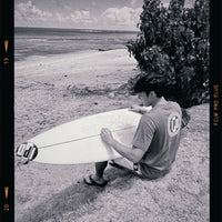 kainalu sitting on the beach wearing an ipd tee, waxing his board and getting ready to surf