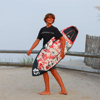 maddox walking in the sand holding his surfboard, turning back and smiling at something behind him looking stoked