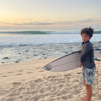 mauri standing on the beach holding his surfboard in front of a beautiful sunset with waves breaking behind him