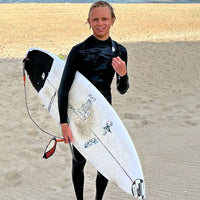 ryan standing on the beach with a wetsuit on carrying his surfboard, smiling, and looking stoked