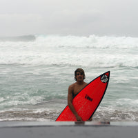 mikel standing just out of the water, holding his surfboard, looking accomplished, with some big waves breaking behind him