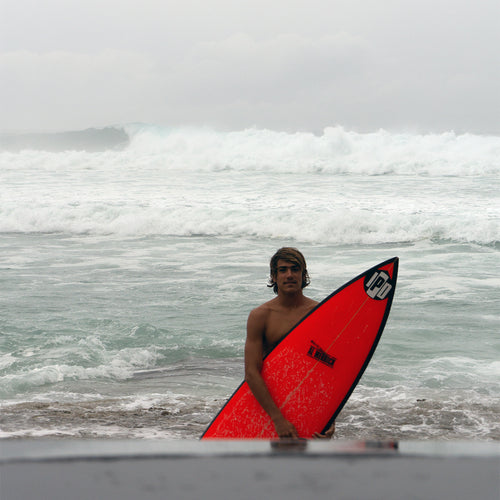 mikel standing just out of the water, holding his surfboard, looking accomplished, with some big waves breaking behind him