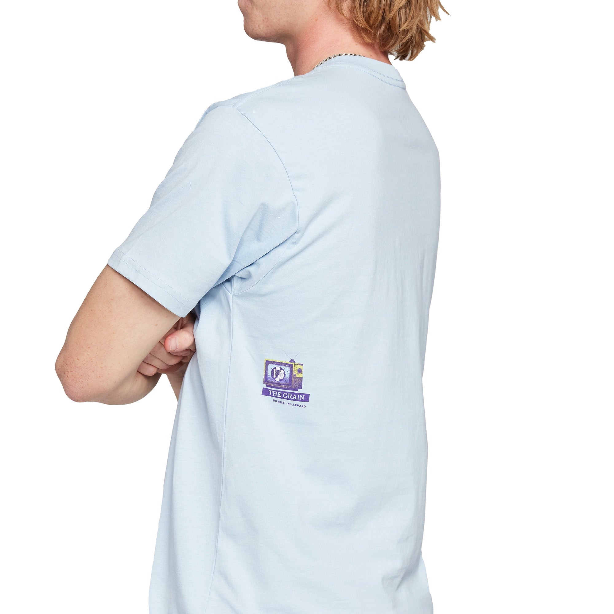 Side view of a light blue short sleeve tee. Just under the armpit is a small I P D logo on the broken screen of an old T V with the words The Grain just below it, and the words No Risk No Reward just below that.