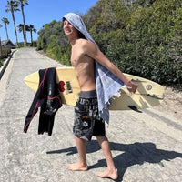 noah walking on a beach path in board shorts holding his surfboard with a towel hanging from his head