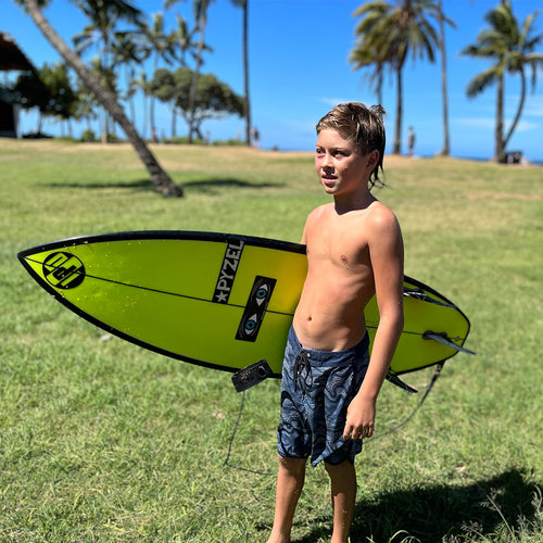 noah standing in the sun on the grass in his boardies with surfboard in hand looking satisfied