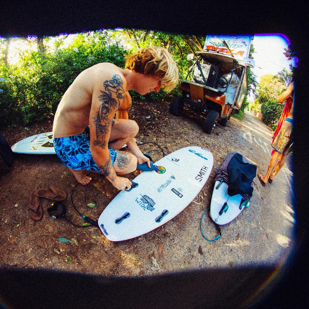 A shirtless surfer is bent over waxing his surfboard while wearing light blue boardshorts with a dark blue, white lined, abstract offset floral print.