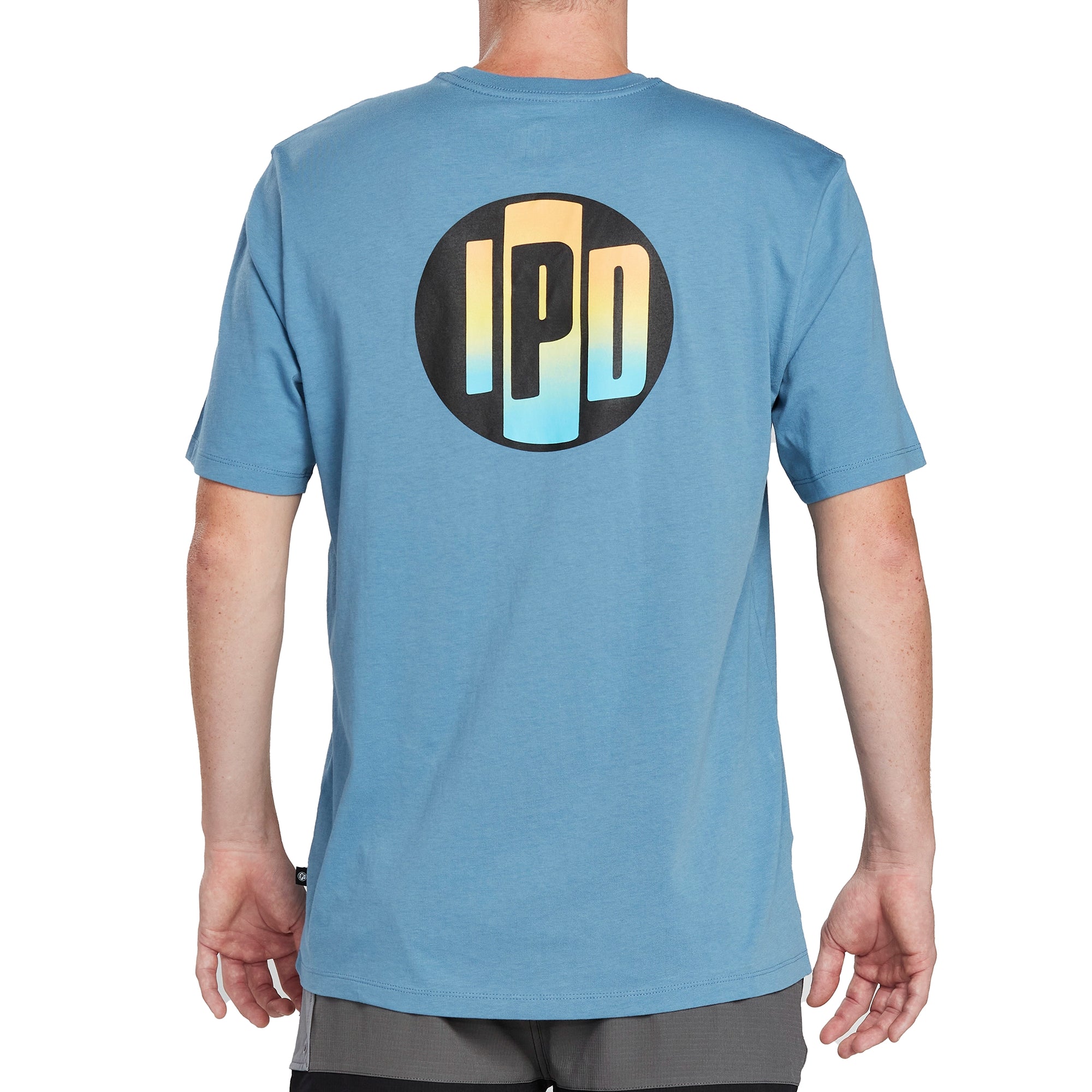 Back of a light blue short sleeve tee with a large print of the I P D logo on the back. The logo is outlined in black with rainbow colors filled in.