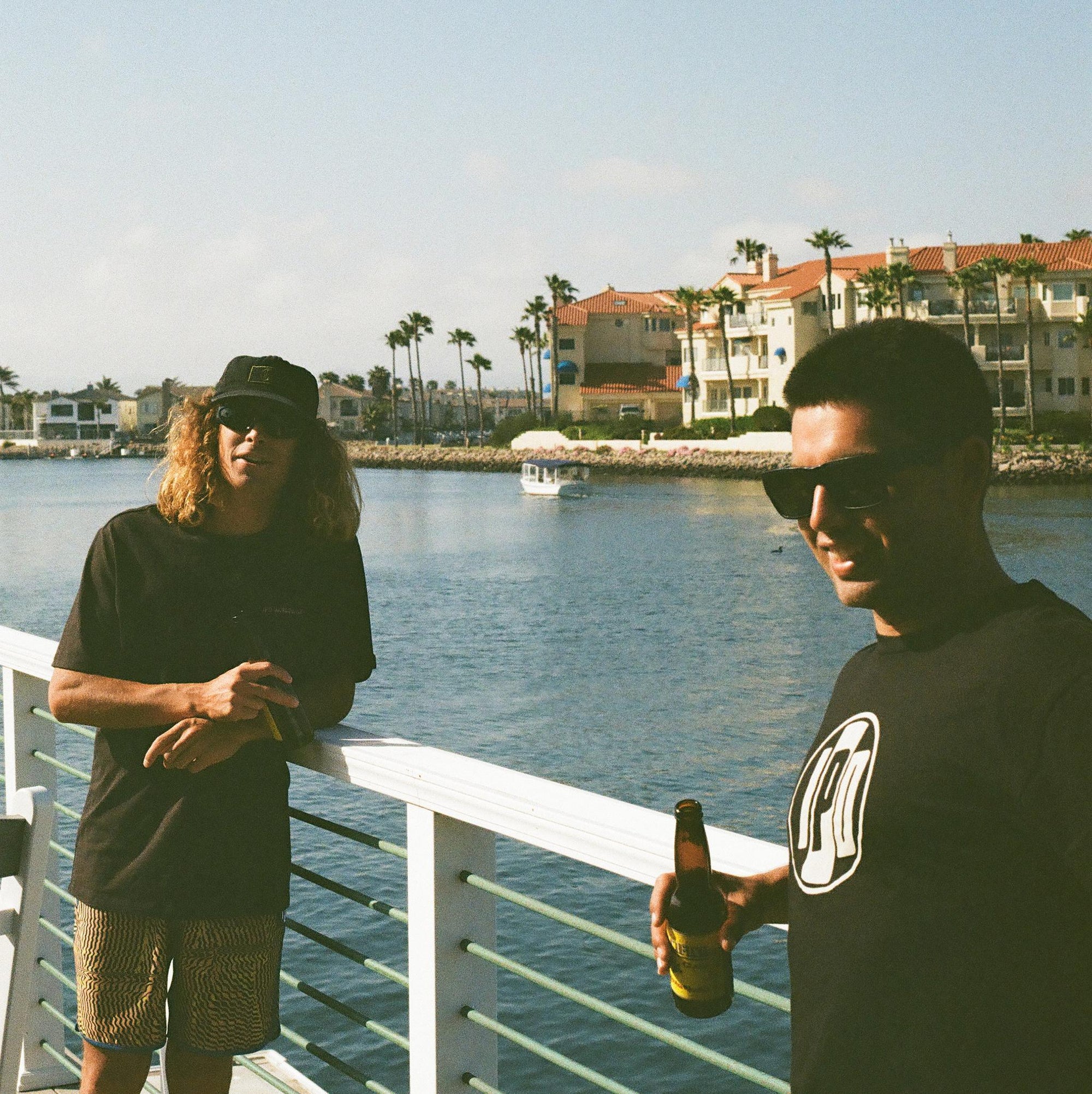 Two men are hanging out on the railing of a dock and drinking beers. One of them is wearing black and yellow abstract striped boardshorts.