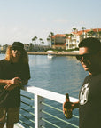 Two men are hanging out on the railing of a dock and drinking beers. One of them is wearing black and yellow abstract striped boardshorts.