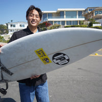 raiki standing outside on the street holding his surfboard with a big smile on his face