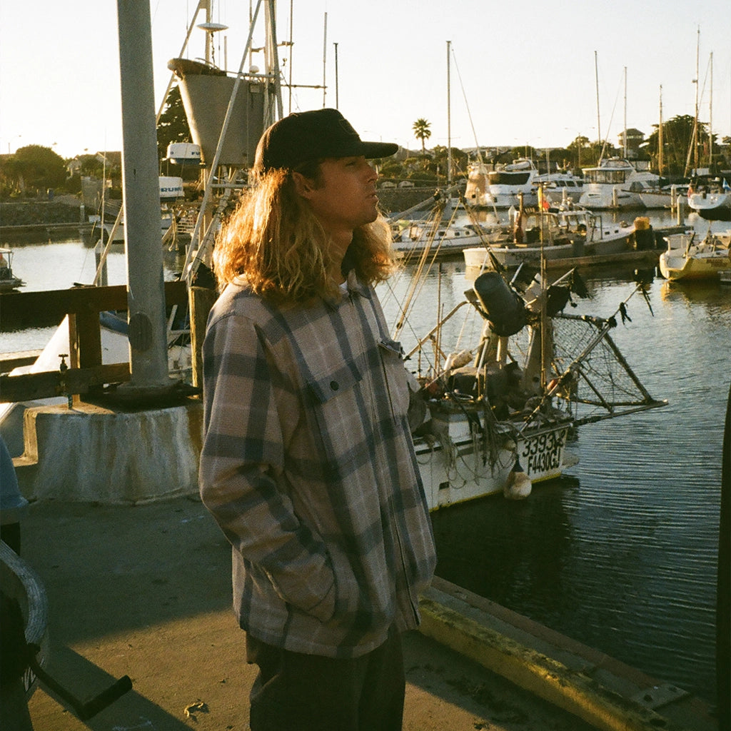 Man standing on a dock in front of a fishing boat wearing a fleece hooded white beige flannel jacket with navy blue stripes.