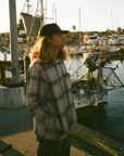 Man standing on a dock in front of a fishing boat wearing a fleece hooded white beige flannel jacket with navy blue stripes.