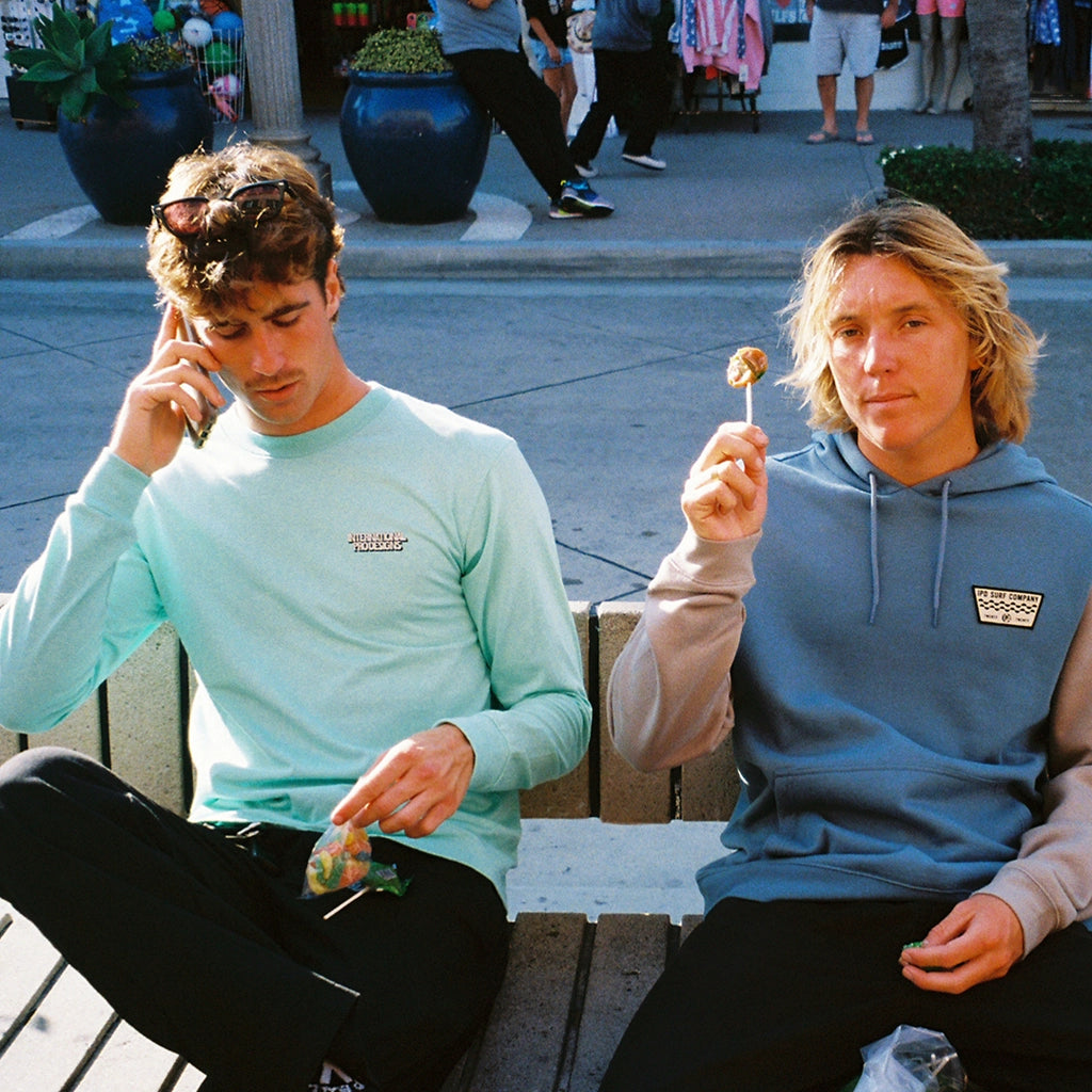 A man sitting on a bench next to his friend wearing a tulum teal long sleeve tee while talking on the phone.