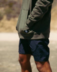 Side view of a man standing outside wearing an olive green hoodie and solid black lightweight boardshorts.