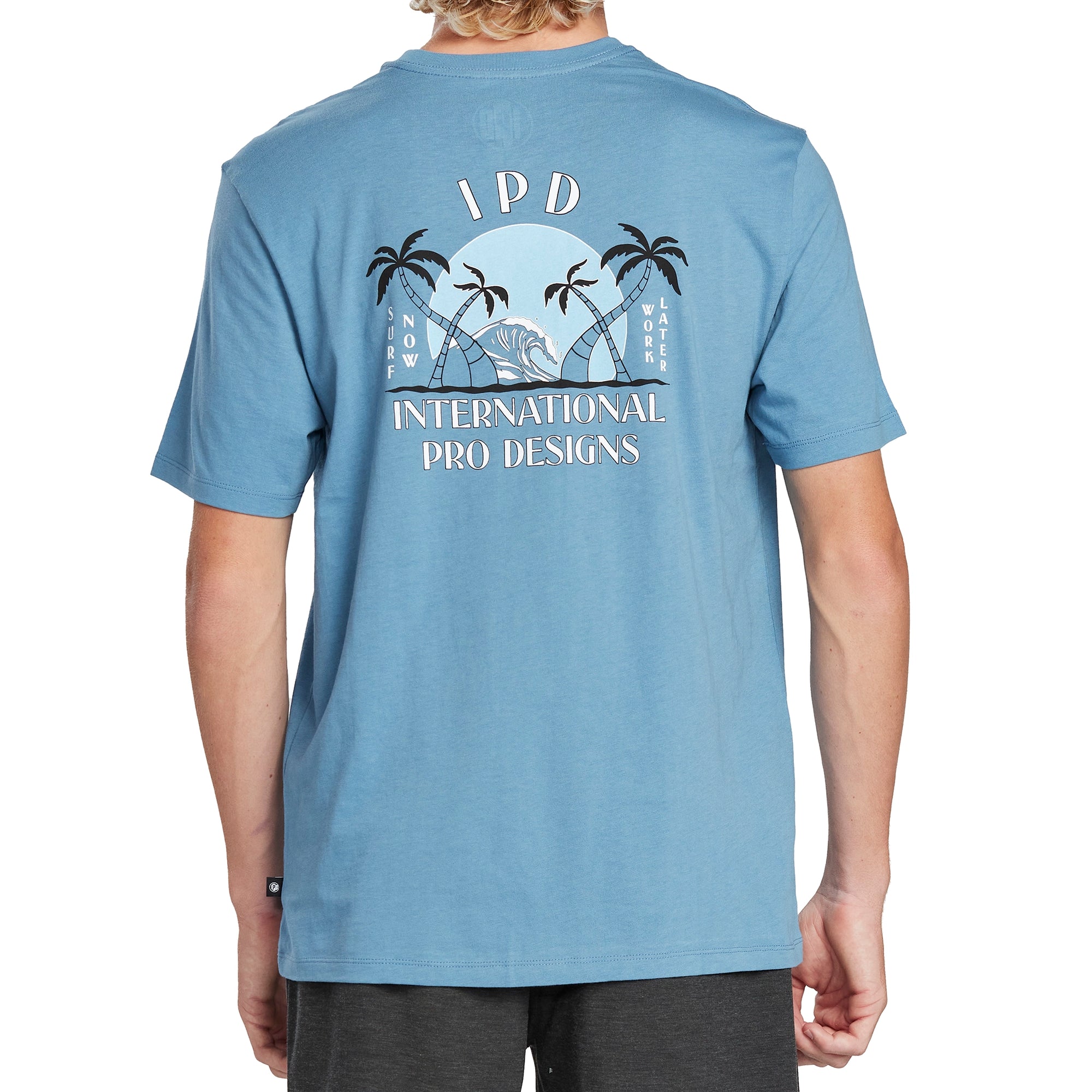 Back of a black short sleeve tee with a large print of a beach with two crossing palm trees on each side and a large breaking wave in the middle. Behind the wave is a large red setting sun. Above the print are the letters I P D and below are the words International Pro Designs. On the left side of the print are the words Surf Now. On the right side the words Work Later.