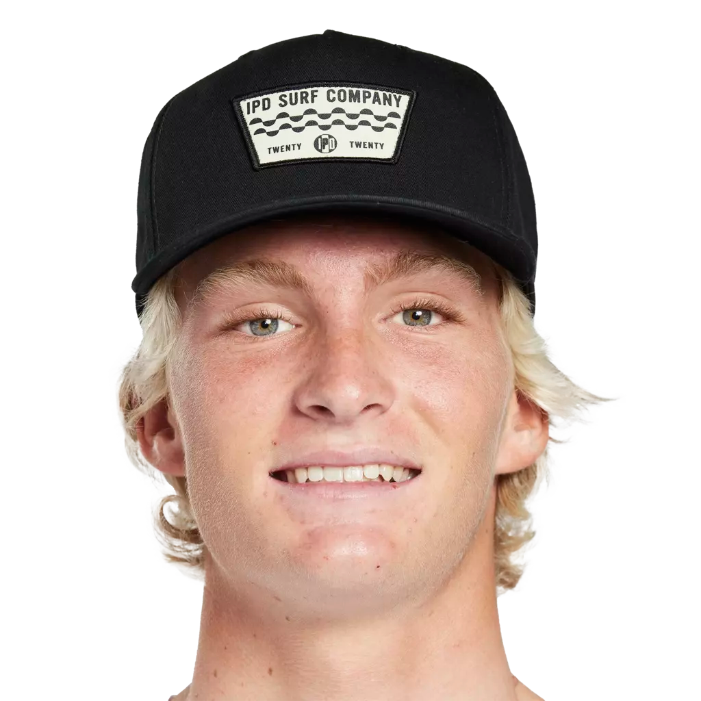 Headshot of a young man wearing a black hat with a white patch on the front. There are two wavy decorative lines running across the middle of the patch. The words I P D Surf Company are above the lines and there&#39;s a small I P D logo below the lines with the word Twenty on each side.