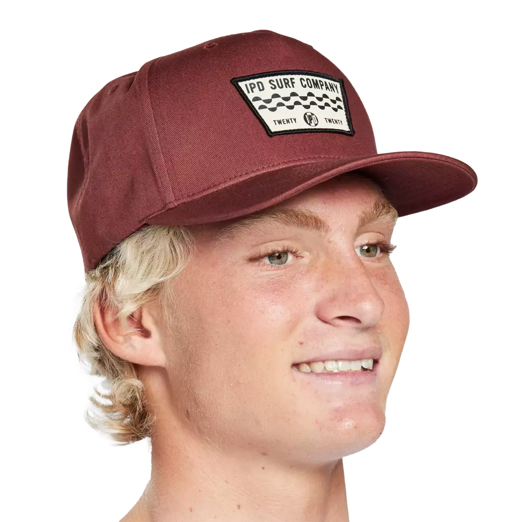 Front side headshot of a young man wearing a burnt red hat with a white patch on the front. There are two wavy decorative lines running across the middle of the patch. The words I P D Surf Company are above the lines and there&#39;s a small I P D logo below the lines with the word Twenty on each side.