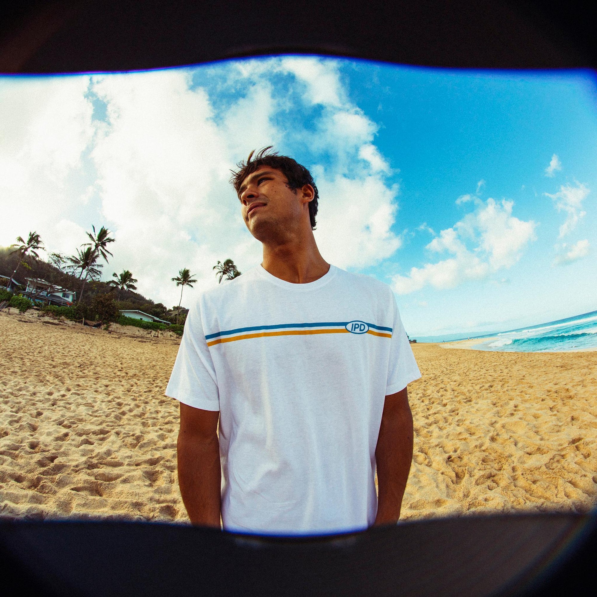 A man standing on the beach looking over to his right, wearing a white short sleeve tee with two stripes across the chest and the letters I P D over the heart in a classic, old school style.