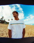 A man standing on the beach looking over to his right, wearing a white short sleeve tee with two stripes across the chest and the letters I P D over the heart in a classic, old school style.