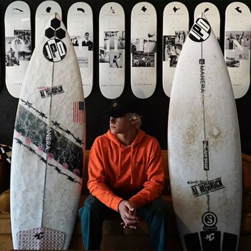 tim sitting down and posing next to two surfboards on either side of him with 8 skateboard decks neatly hanging on the wall behind him